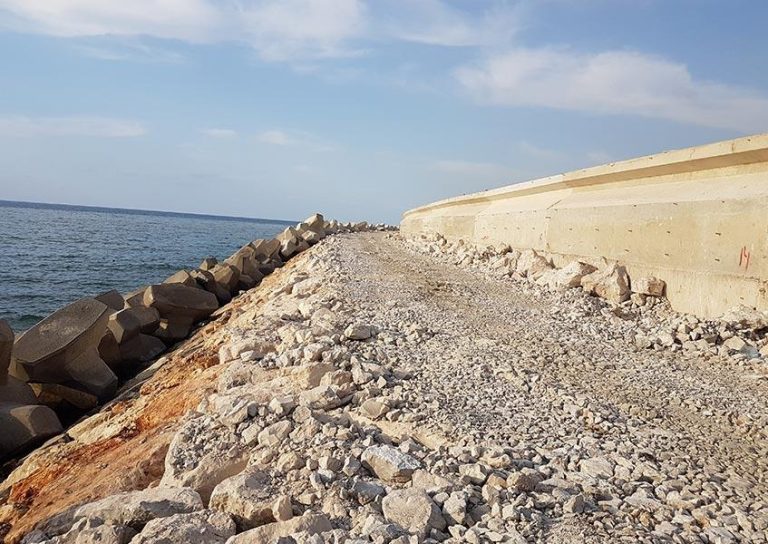 A rocky path runs parallel to the sea, bordered on one side by large concrete blocks and on the other by a high concrete wall. The sky is partly cloudy, and the sea appears calm under the sunlight.