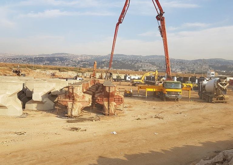 A construction site with various heavy machinery, including cranes, cement trucks, and diggers, operating. Large concrete blocks and metal structures are visible on the ground. In the background, there are distant hills and a partly cloudy sky.