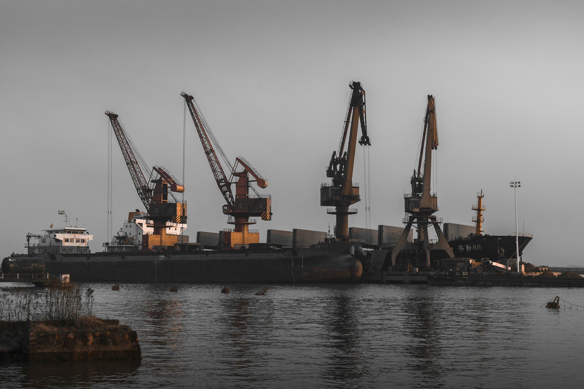 A harbor scene at dusk showing several large cranes unloading a ship docked at a port. The sky is overcast, and the lighting is dim, giving the image a moody atmosphere. The water in the foreground reflects the muted colors of the sky.