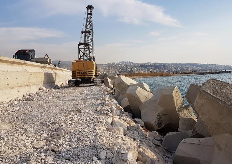 A construction crane on a rocky path beside large concrete blocks along the coastline. The sea is on the right, and a cityscape is visible in the background under a clear sky with a few clouds. A truck is parked to the left near the crane.