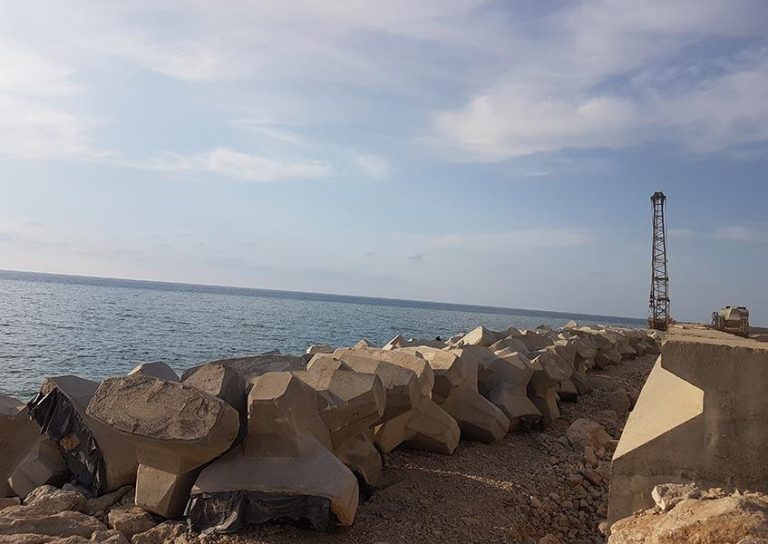 A coastal scene with large, geometric concrete blocks arranged along the shore. The sea stretches out under a partly cloudy sky. On the right, a crane or derrick stands on the rocky terrain near the water.