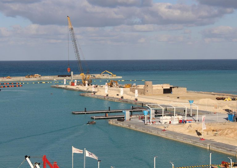 A construction site along a seaside harbor with turquoise water. The site includes a crane, construction equipment, and several unfinished structures. The background features a calm blue sea and a partly cloudy sky.