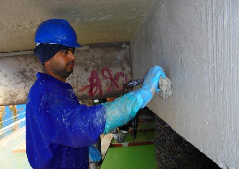 A worker in a blue helmet and jumpsuit applies a grey substance to a large surface with a brush. He is wearing gloves and concentrating on the task. Graffiti can be seen on the wall behind him.
