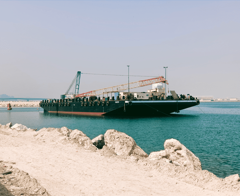 A large barge with a crane on its deck is moored close to a rocky shoreline in calm, teal water. The sky is clear and light blue. Some distant structures and another vessel are visible against the horizon.