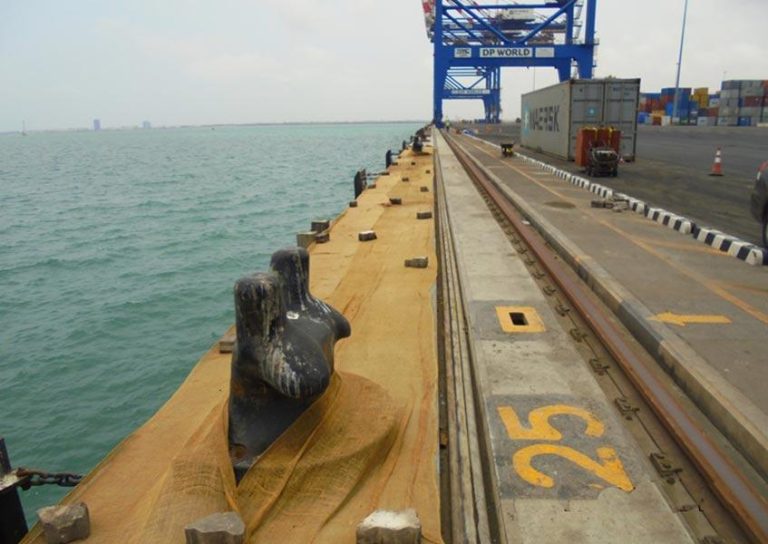 A seaside shipping port with a large crane marked "DP World" in the background, shipping containers stacked to the right side, and the number "25" prominently marked on the concrete surface near the water.