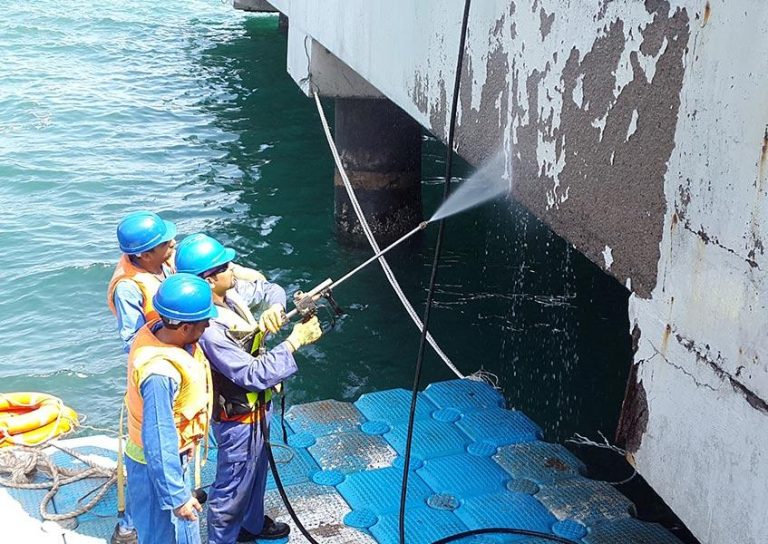 Three workers wearing blue hard hats, safety vests, and safety glasses are using a high-pressure water jet to clean or remove old paint from a concrete surface near a body of water. They are standing on floating platforms with visible ropes for safety.