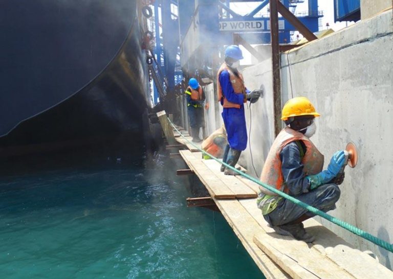 Workers in safety gear, including helmets and vests, perform maintenance tasks on a large, docked ship. They are using tools to grind and polish surfaces while standing on narrow wooden scaffolding suspended over water. Industrial equipment is visible in the background.