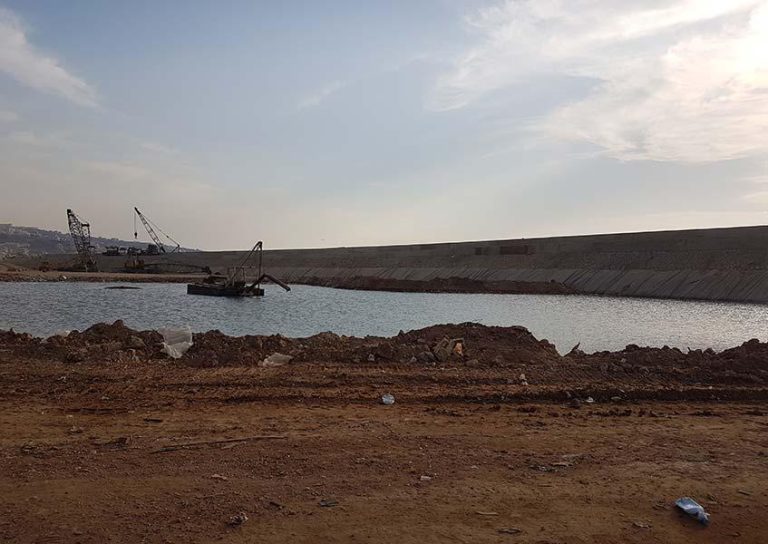 A construction site near a body of water, with two cranes in the background. The ground is mostly dirt with some construction debris scattered around. The sky is partly cloudy, and a concrete structure runs parallel to the water.