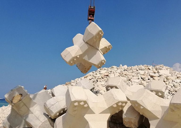 A large, geometric concrete block is being lifted by a crane against a clear blue sky. Several similar blocks are piled on a rocky shore in the background. One person is visible among the blocks.