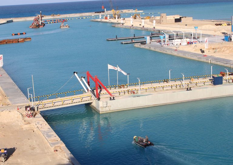 A view of a calm coastal construction site with clear blue water. A small boat with two people is visible in the foreground, while cranes and other construction machinery are working in the background. A bridge spans across part of the water.