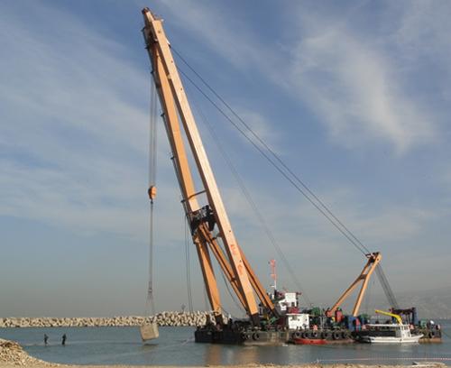 A large floating crane is positioned in the water near a rocky shoreline under a partly cloudy sky. The crane is hoisting a concrete block with extended cables. Several workers and small boats are nearby, suggesting construction or infrastructure work.