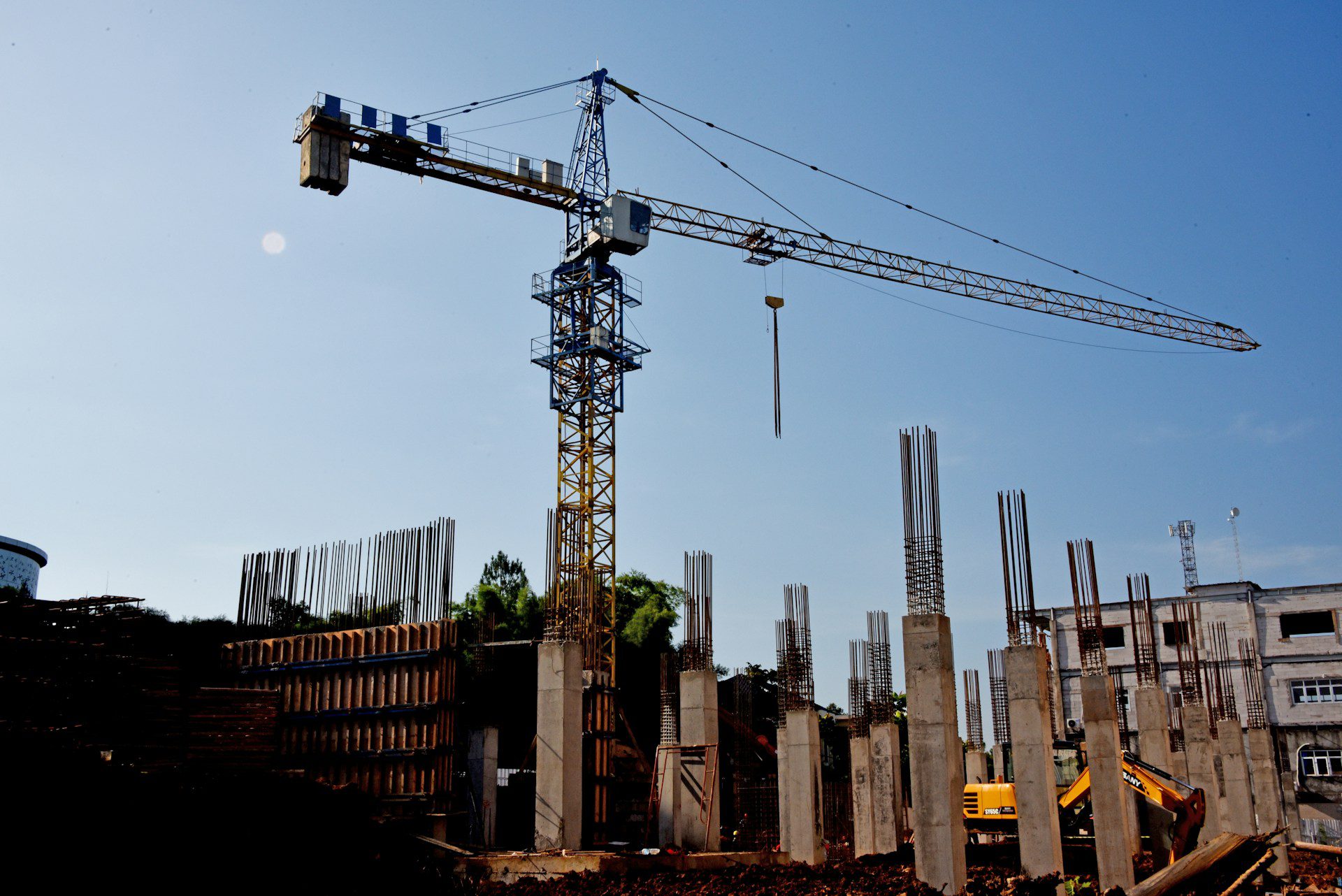 A construction site with a tall yellow and blue crane towering over partially built structures. Steel rebar protrudes from concrete columns, and there is a yellow excavation vehicle. The sky is clear and blue, providing a bright backdrop.