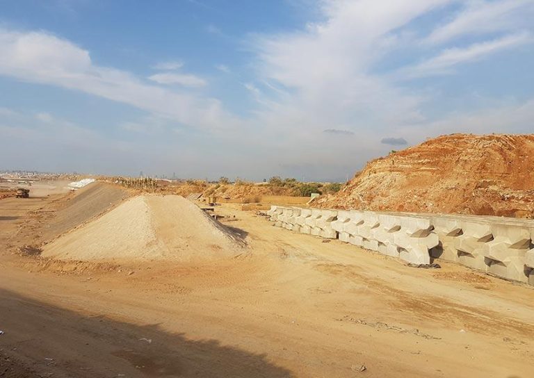 Construction site with mounds of sand, a row of large concrete blocks, and a partially excavated hill. The sky is blue with light clouds. Scattered machinery and tools are visible in the background. Sparse vegetation can be seen in the distance.