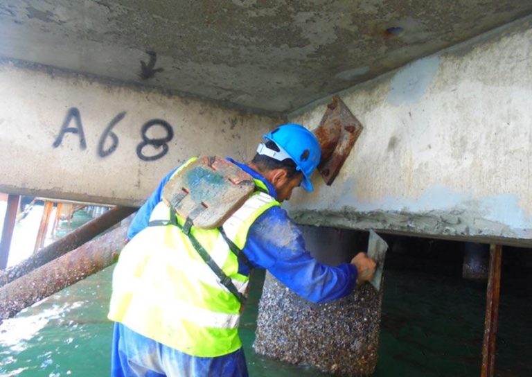 A construction worker in a blue hard hat, gloves, and a high-visibility vest is repairing underwater concrete foundations. The worker is applying material to a beam marked "A68." The structure is partially submerged in water.