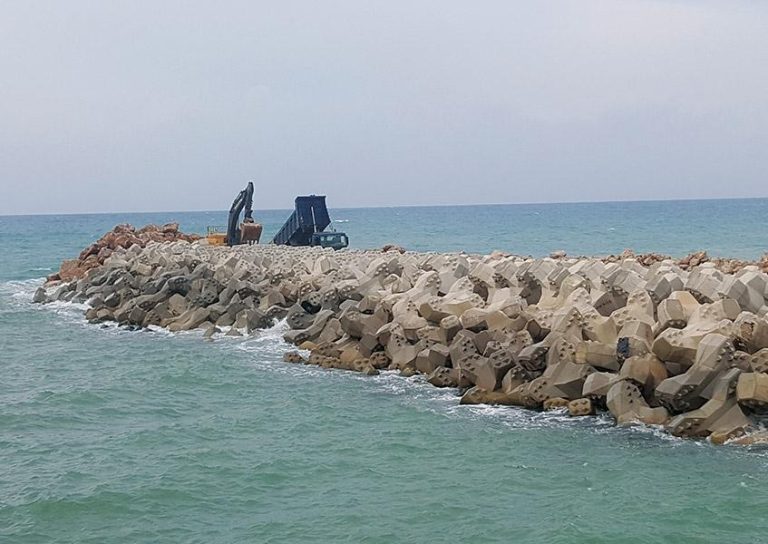 A construction site on a breakwater extends into the sea. Two pieces of machinery, a blue dump truck and an excavator, are positioned on the breakwater, surrounded by large interlocking concrete blocks. Waves gently hit the structure. The sky is cloudy.