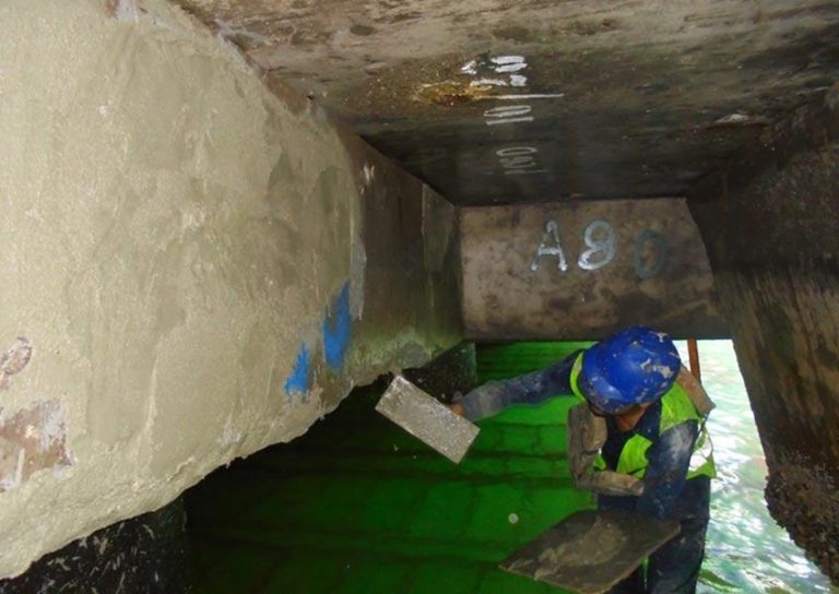 A worker wearing a hard hat and safety vest is applying cement or a similar substance to the underside of a concrete structure. The area is marked with "A80" and is supported over a body of green-colored water.