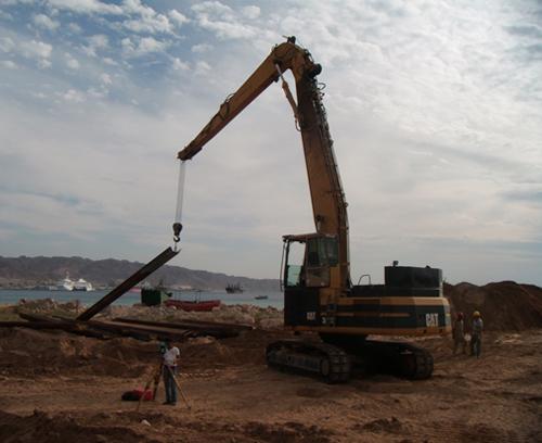 An excavator is lifting a long, metal beam at a construction site near a body of water. The sky is partly cloudy, and several boats are visible in the background. Two workers are on-site, and one is taking a photo.