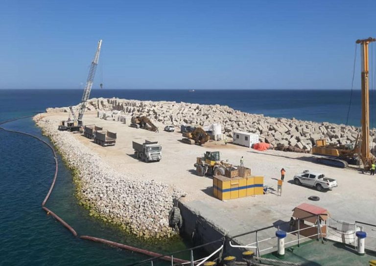 A construction site on a coastal pier with various vehicles and machinery. Workers are present, and concrete blocks are stacked nearby. The site extends into calm blue water under a clear sky and is part of a larger body of water, possibly an ocean or sea.