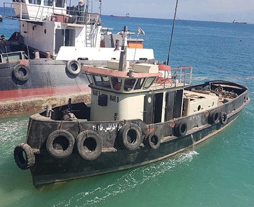 A small towing vessel is anchored near a larger ship. The boat is equipped with tires along its sides, presumably serving as fenders. The vessel's name, "Mar 9," is visible on the front. The sea around them is clear and blue, indicating a sunny day.