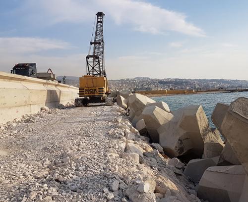 A large yellow construction crane operates on a rocky coastal path beside the sea. The path is lined with concrete blocks and heavy machinery is visible. In the background, a cityscape stretches along the horizon under a partly cloudy sky.