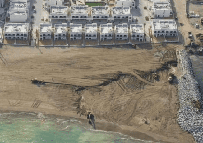 Aerial view of a coastal construction site with several bulldozers and excavators working on the sandy shore. Nearby are rows of white buildings, suggesting a residential area, with the ocean visible at the bottom of the image.
