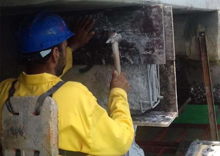 A construction worker wearing a blue helmet and a yellow shirt is using a hammer to work on a concrete structure. The worker is standing under a large slab, focusing on the task at hand. Tools and materials are visible nearby.