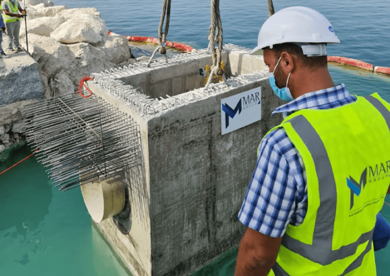 A construction worker wearing a hard hat and high-visibility vest oversees the lowering of a concrete structure, reinforced with metal bars, into a body of water using a crane. Other workers and rock structures are visible in the background.