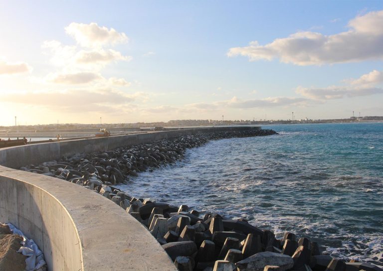 A curved concrete seawall extends along a coast, with large wave breakers lining the shore to prevent erosion. The calm sea stretches to the horizon under a partly cloudy sky, and sunlight illuminates the distant land in the background.