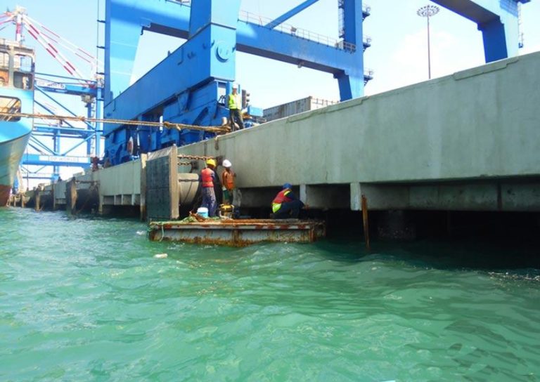 Workers in safety gear stand on a floating platform, performing maintenance on a concrete structure by the water. Large blue cranes loom in the background, indicating an industrial or port environment. The water is a clear blue-green.
